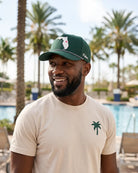 A smiling man wearing a green baseball cap with a white Florida state graphic and a beige t-shirt with a green palm tree logo, standing outdoors in front of a swimming pool with palm trees.
