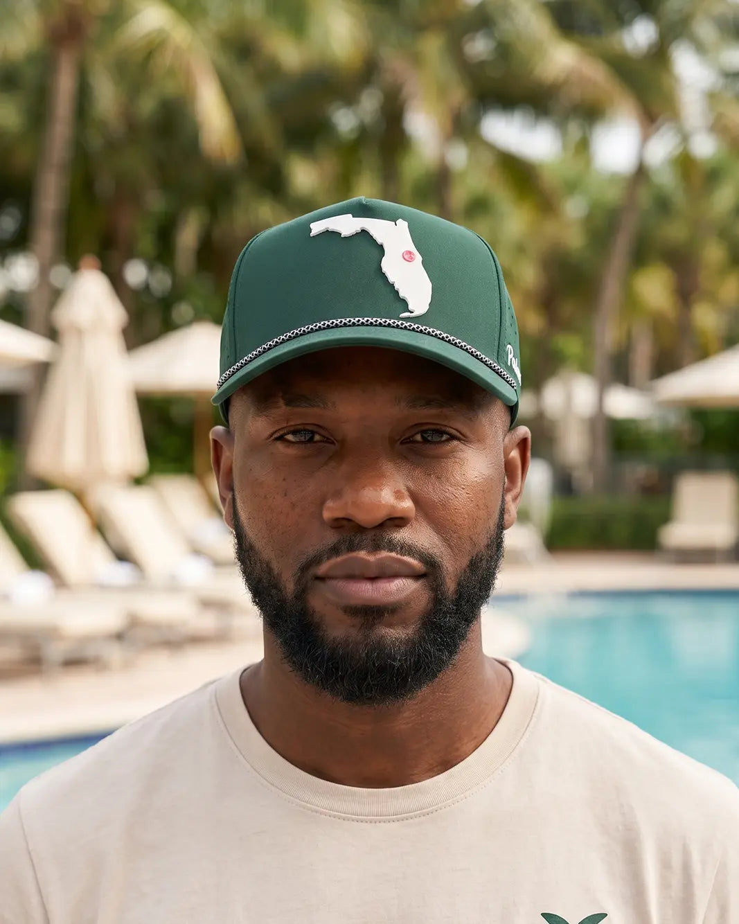 A man wearing a green baseball cap with a white Florida state silhouette and a small red palm tree, standing in front of a swimming pool with palm trees in the background.