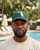 A man wearing a green baseball cap with a white Florida state silhouette and a small red palm tree, standing in front of a swimming pool with palm trees in the background.