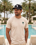 A man wearing a beige crewneck t-shirt with a green palm tree logo on the left chest, paired with a black baseball cap, standing in front of a swimming pool with palm trees in the background.