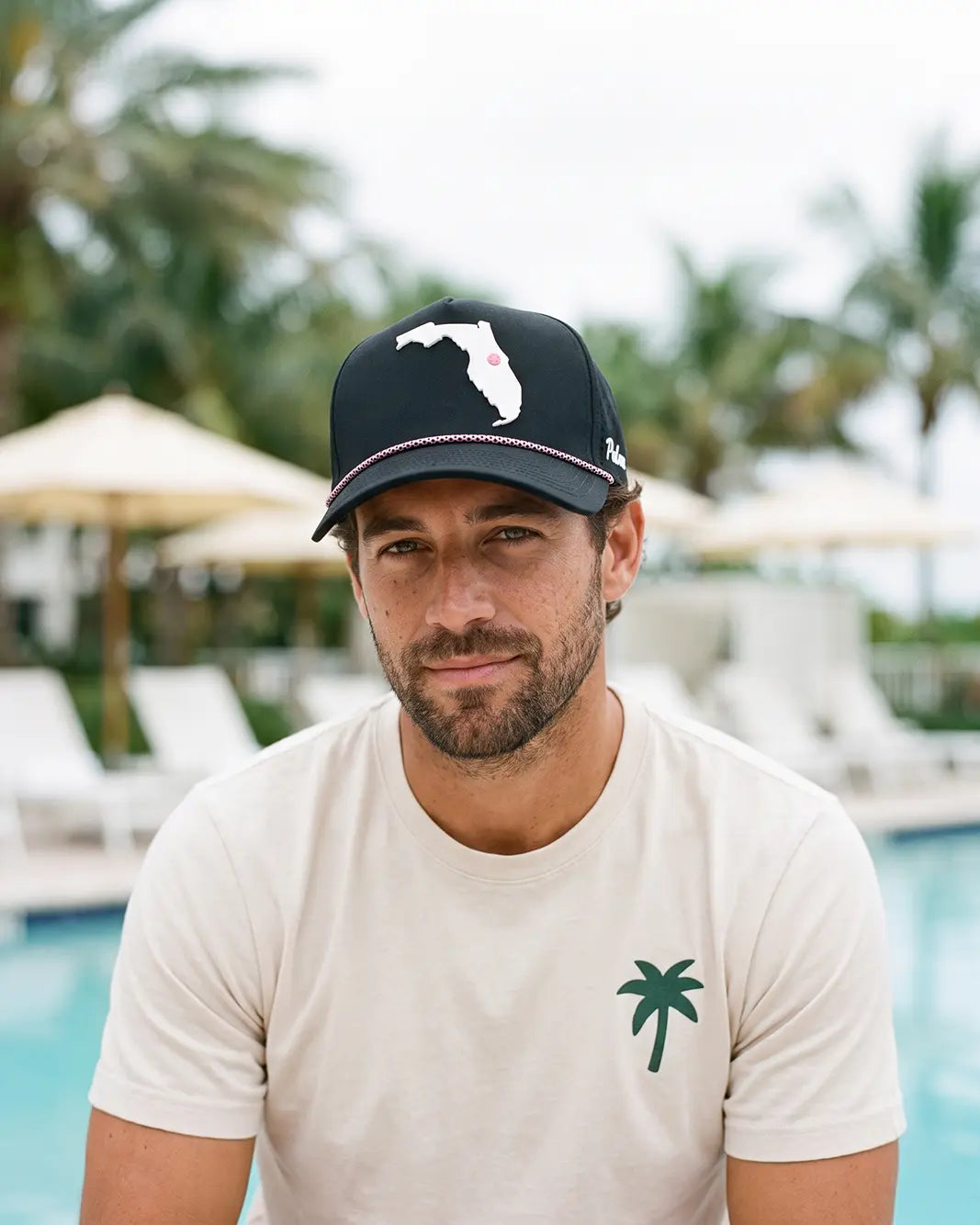 A man wearing a cream-colored t-shirt with a green palm tree logo and a black baseball cap with a white Florida state silhouette, posed in front of a pool with palm trees and lounge chairs in the background.