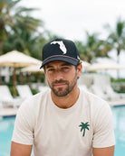 A man wearing a cream-colored t-shirt with a green palm tree logo and a black baseball cap with a white Florida state silhouette, posed in front of a pool with palm trees and lounge chairs in the background.