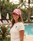 A woman smiling outdoors by a swimming pool, wearing a pink baseball cap with a white Florida state outline and a white t-shirt with a small green palm tree logo.