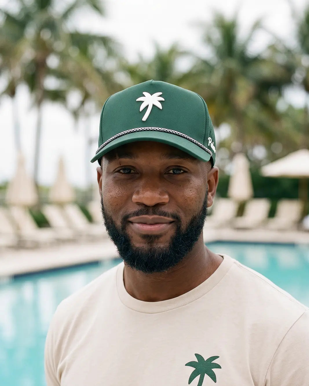 A man wearing a green baseball cap with a white palm tree logo and a beige t-shirt, standing in front of a blurred swimming pool and palm trees.