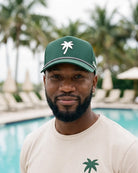 A man wearing a green baseball cap with a white palm tree logo and a beige t-shirt, standing in front of a blurred swimming pool and palm trees.