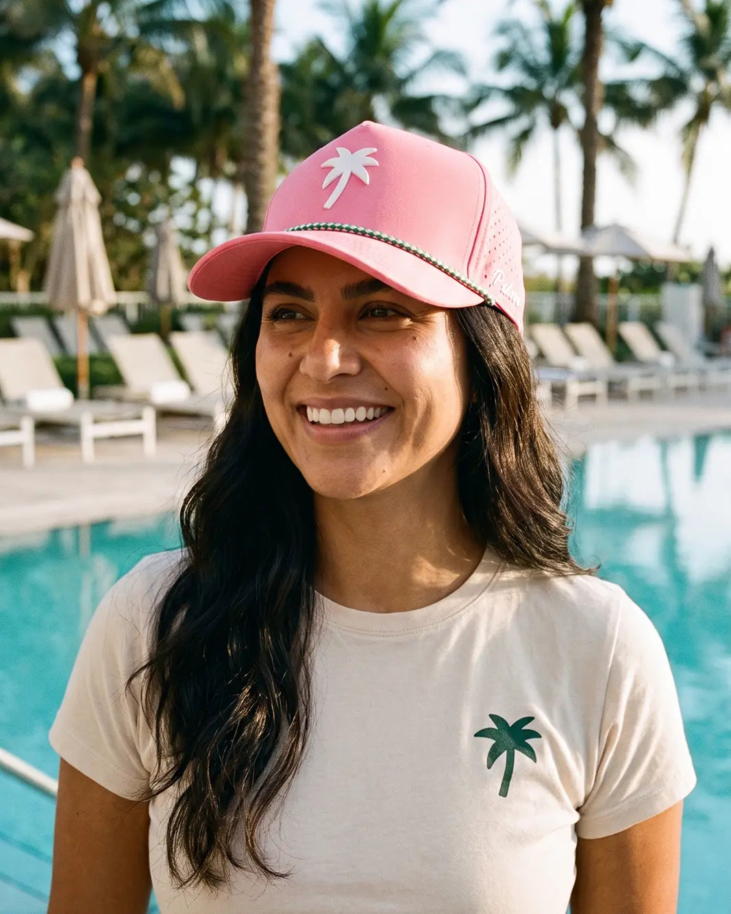 A smiling woman standing by a swimming pool wearing a pink trucker-style cap with a white palm tree graphic and a cream T-shirt with a green palm tree logo. Palm trees and lounge chairs are visible in the background.