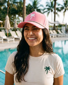 A smiling woman standing by a swimming pool wearing a pink trucker-style cap with a white palm tree graphic and a cream T-shirt with a green palm tree logo. Palm trees and lounge chairs are visible in the background.