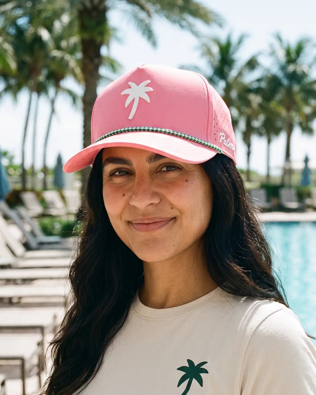 A woman wearing a pink baseball cap with a white palm tree logo and a green and white beaded trim, standing outdoors near a swimming pool with palm trees in the background.