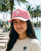 A woman wearing a pink baseball cap with a white palm tree logo and a green and white beaded trim, standing outdoors near a swimming pool with palm trees in the background.