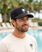 A man wearing a black baseball cap with a white palm tree logo and a cream t-shirt, photographed outdoors in front of a swimming pool with palm trees in the background.