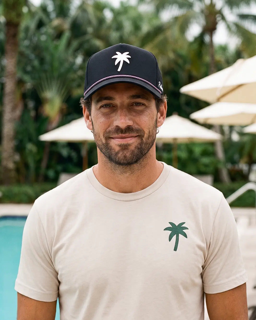 A man wearing a beige t-shirt with a green palm tree logo and a navy blue baseball cap with a white palm tree logo, standing in front of a pool with palm trees and umbrellas in the background.