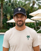 A man wearing a beige t-shirt with a green palm tree logo and a navy blue baseball cap with a white palm tree logo, standing in front of a pool with palm trees and umbrellas in the background.