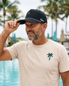 A smiling man wearing a beige t-shirt and a black trucker hat with a palm tree logo, standing by a swimming pool with palm trees in the background.