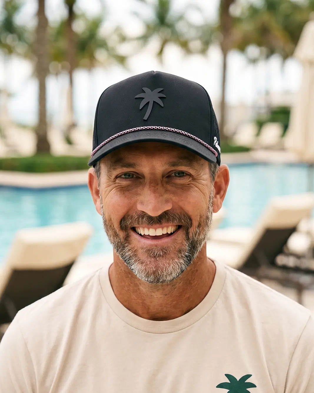 A smiling man wearing a black baseball cap with a dark gray palm tree logo and a beige t-shirt, photographed outdoors with a swimming pool and palm trees in the background.