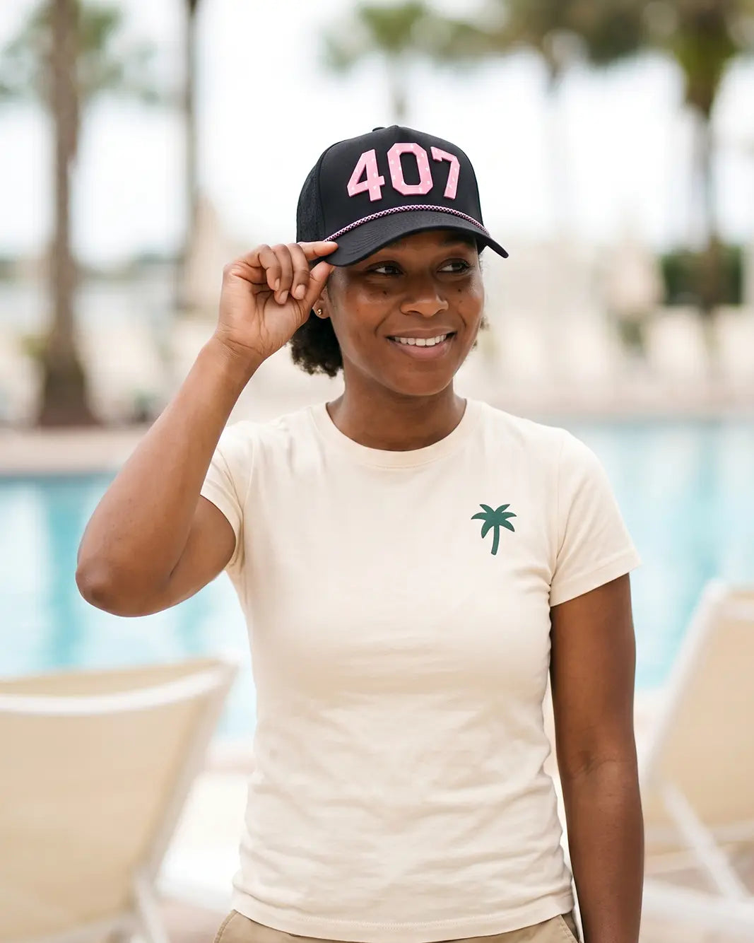 The 407 Signature Hat (Pink on Black) worn on-model on a pool deck, hand touching the brim, three-quarter view: pink 407 on a black crown.