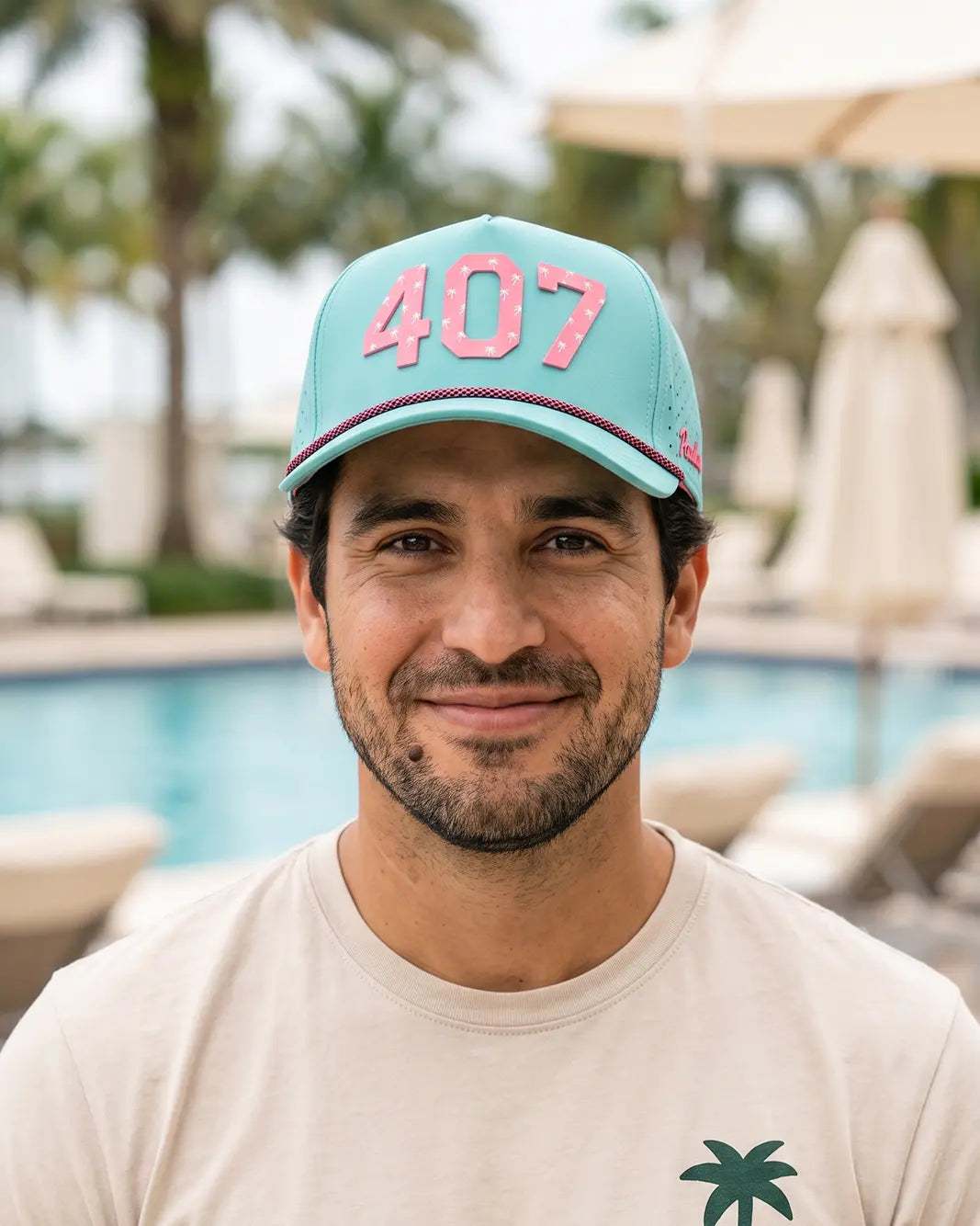A man wearing a light blue baseball cap with pink '407' lettering and a beige t-shirt, photographed outdoors in front of a swimming pool with palm trees and lounge chairs.