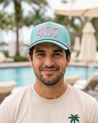 A man wearing a light blue baseball cap with pink '407' lettering and a beige t-shirt, photographed outdoors in front of a swimming pool with palm trees and lounge chairs.