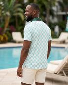 A man standing by a swimming pool wearing a mint green and white checkered short-sleeve polo shirt with a dark green collar, paired with cream-colored shorts.