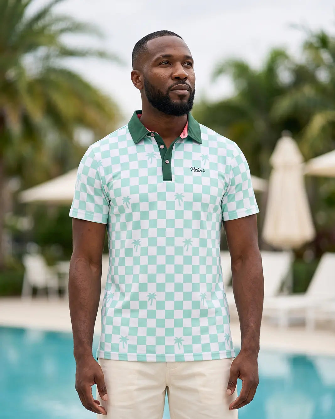 A man wearing a mint green and white checkered polo shirt with a green collar, standing in front of a pool with palm trees in the background. The brand name 'Palmers' is embroidered on the chest.