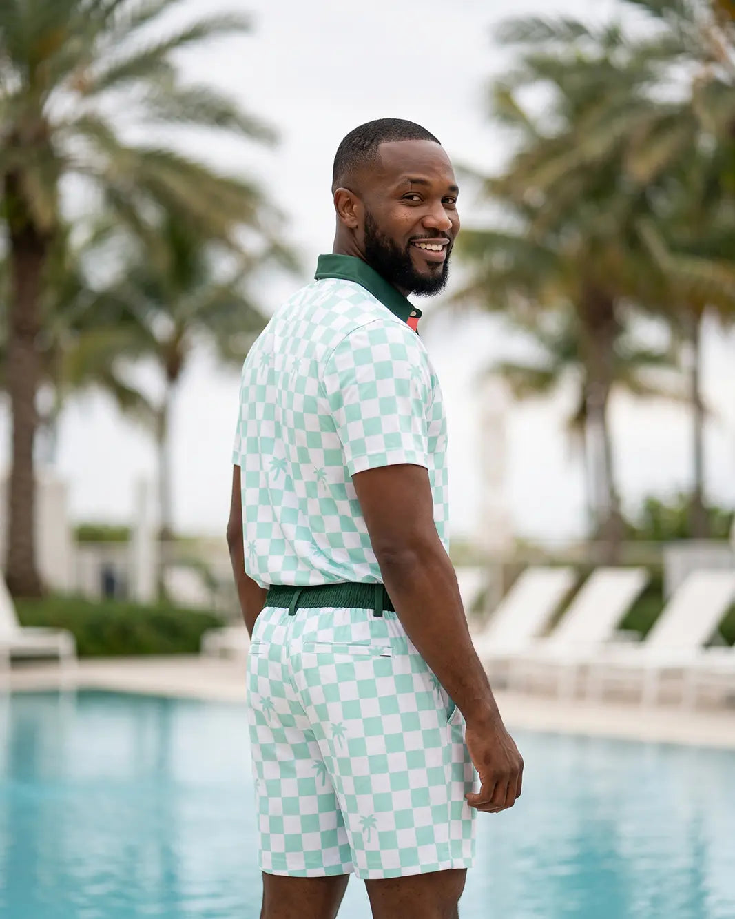 A man wearing a mint green and white checkered polo shirt and matching shorts stands by a swimming pool with palm trees in the background.