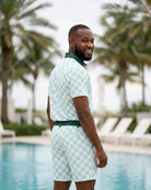 A man wearing a mint green and white checkered polo shirt and matching shorts stands by a swimming pool with palm trees in the background.