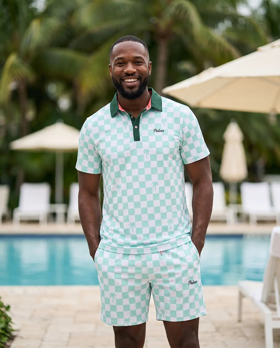 A man wearing a light green and white checkered polo shirt and matching shorts stands in front of a swimming pool with palm trees and umbrellas in the background. The brand name 'Paisley' is visible on the chest of the polo shirt.