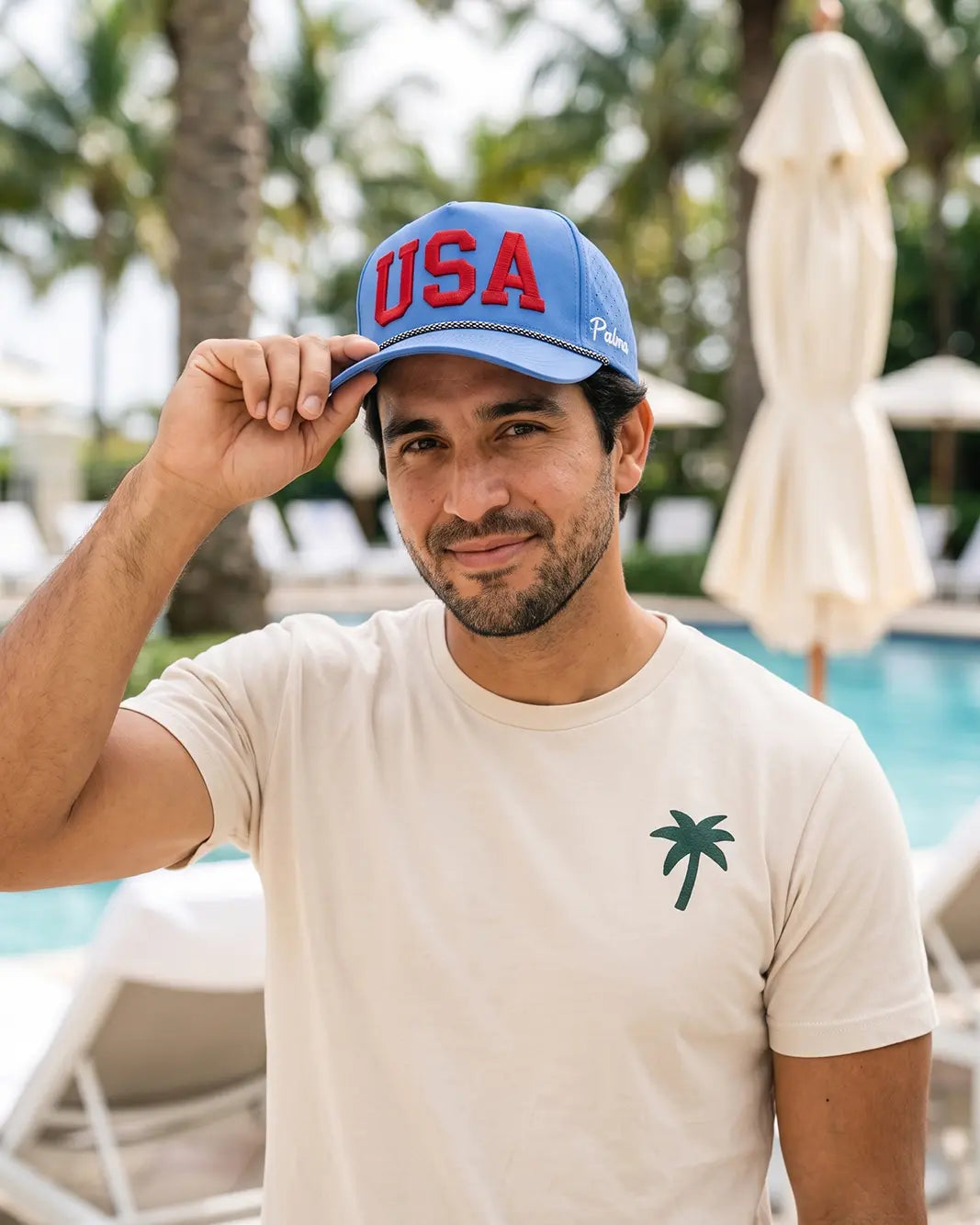 A man wearing a light beige t-shirt with a small green palm tree logo and a blue 'USA' trucker hat, posing in front of a pool with palm trees in the background.