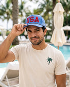 A man wearing a light beige t-shirt with a small green palm tree logo and a blue 'USA' trucker hat, posing in front of a pool with palm trees in the background.