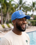 The 239 Signature Hat (Blue) worn on-model poolside, side view with the perforated panel and palm badge, white 239 above the brim.