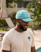 The 772 Signature Hat (Teal) worn on-model by the pool, head tilted down, side view: perforated panel, pink palm badge, white 772 and pink rope.