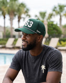 The 863 Signature Hat (Green) worn on-model, seated by the pool, three-quarter view: white 863 on a dark green crown, perforated panel, white Palmr script.