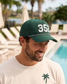 The 352 Signature Hat (Green) worn on-model by the pool, head tilted down, three-quarter view: white 352 with palm print on a dark green crown, perforated panel.