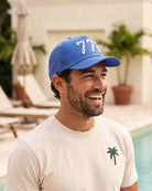 The 772 Signature Hat (Blue) worn on-model in a courtyard, laughing, three-quarter view: white 772 on a blue crown, perforated panel, black and white rope.