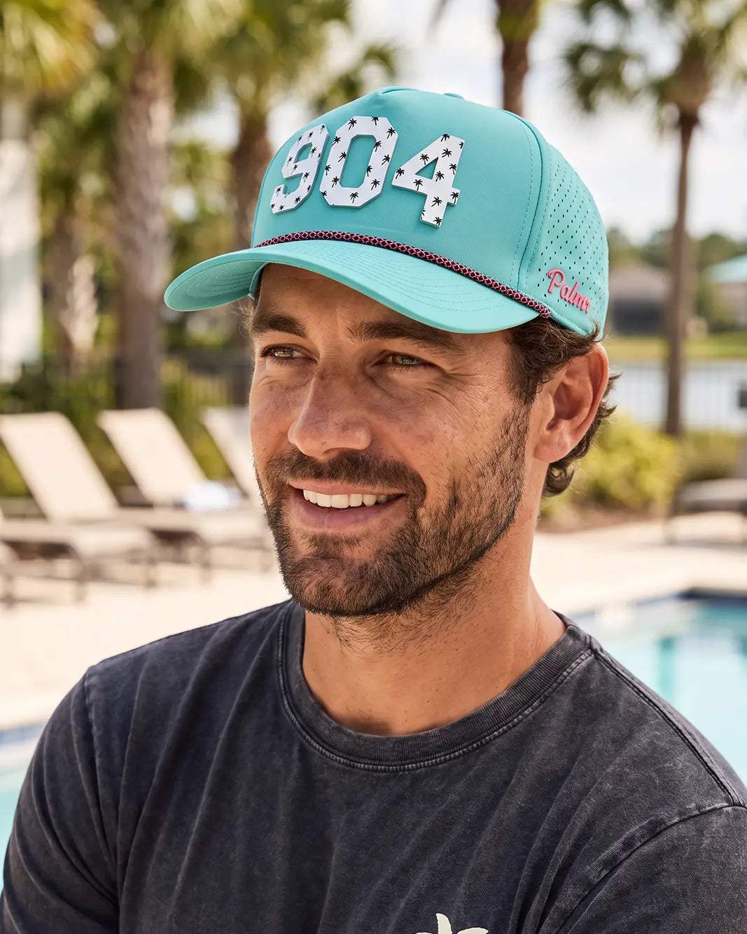 The 904 Signature Hat (Teal) worn on-model by the pool, smiling, three-quarter view: white 904 with palm print on a teal crown, pink rope, pink Palmr script.