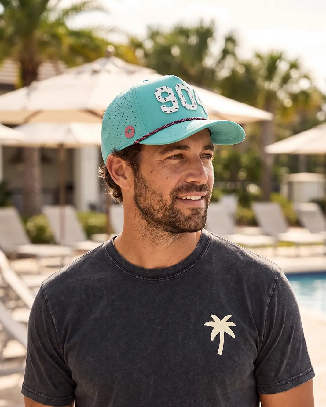 The 904 Signature Hat (Teal) worn on-model poolside, side profile: perforated panel, pink palm badge, white 904 and pink rope on a teal crown.