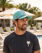 The 904 Signature Hat (Teal) worn on-model poolside, side profile: perforated panel, pink palm badge, white 904 and pink rope on a teal crown.
