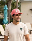 The 352 Signature Hat (Pink) worn on-model on a pool deck, three-quarter view: white 352 with palm print on a pink crown, green rope.