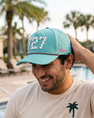 The 727 Signature Hat (Teal) worn on-model poolside, hand on the back of the hat, head down: white 727 on a teal crown, pink rope, pink Palmr script.