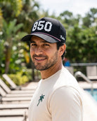The 850 Signature Hat (Black) worn on-model poolside, looking back over the shoulder: white 850 with palm print on a black crown, pink rope, Palmr script.
