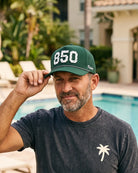 The 850 Signature Hat (Green) worn on-model poolside, hand on the brim: white 850 with palm print on a dark green crown, pink rope, Palmr script.