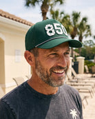 The 850 Signature Hat (Green) worn on-model by the pool, smiling, three-quarter view: white 850 on a dark green crown, perforated panel, pink rope.