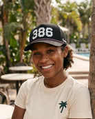 The 386 Signature Hat (Black) worn on-model poolside, front three-quarter view: white 386 with palm print on a black crown, pink rope, Palmr script.