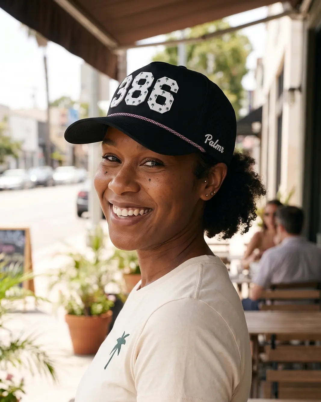The 386 Signature Hat (Black) worn on-model on a cafe patio, looking back over the shoulder: white 386 on a black crown with pink rope and Palmr script.