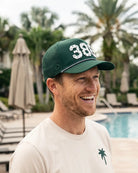 The 386 Signature Hat (Green) worn on-model by the pool, side view laughing: perforated panel, white 386 and pink rope on a dark green crown.