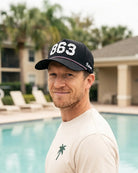 The 863 Signature Hat (Black) worn on-model by the pool, looking back over the shoulder: white 863 on a black crown with pink rope and Palmr script.