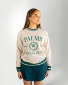 Woman wearing a cream-colored sweatshirt with 'Palmr Social Club' branding on a light gray background