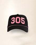 A black trucker-style baseball cap featuring the number '305' in pink with a white star pattern on the front, and a thin pink chain detail across the brim.