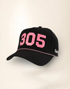 A black baseball cap featuring the number '305' in pink with white polka dots on the front, a pink chain detail across the visor, and white script text on the side, set against a plain off-white background.