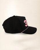 A black baseball cap with a perforated front panel, featuring a pink number '3' on the side and a pink-and-white dotted line detail along the edge of the brim, set against a plain light beige background.