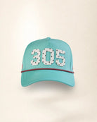 A turquoise baseball cap with '305' embroidered in white with small black star details, accented by a red and black polka dot line across the front, set against a plain cream background.